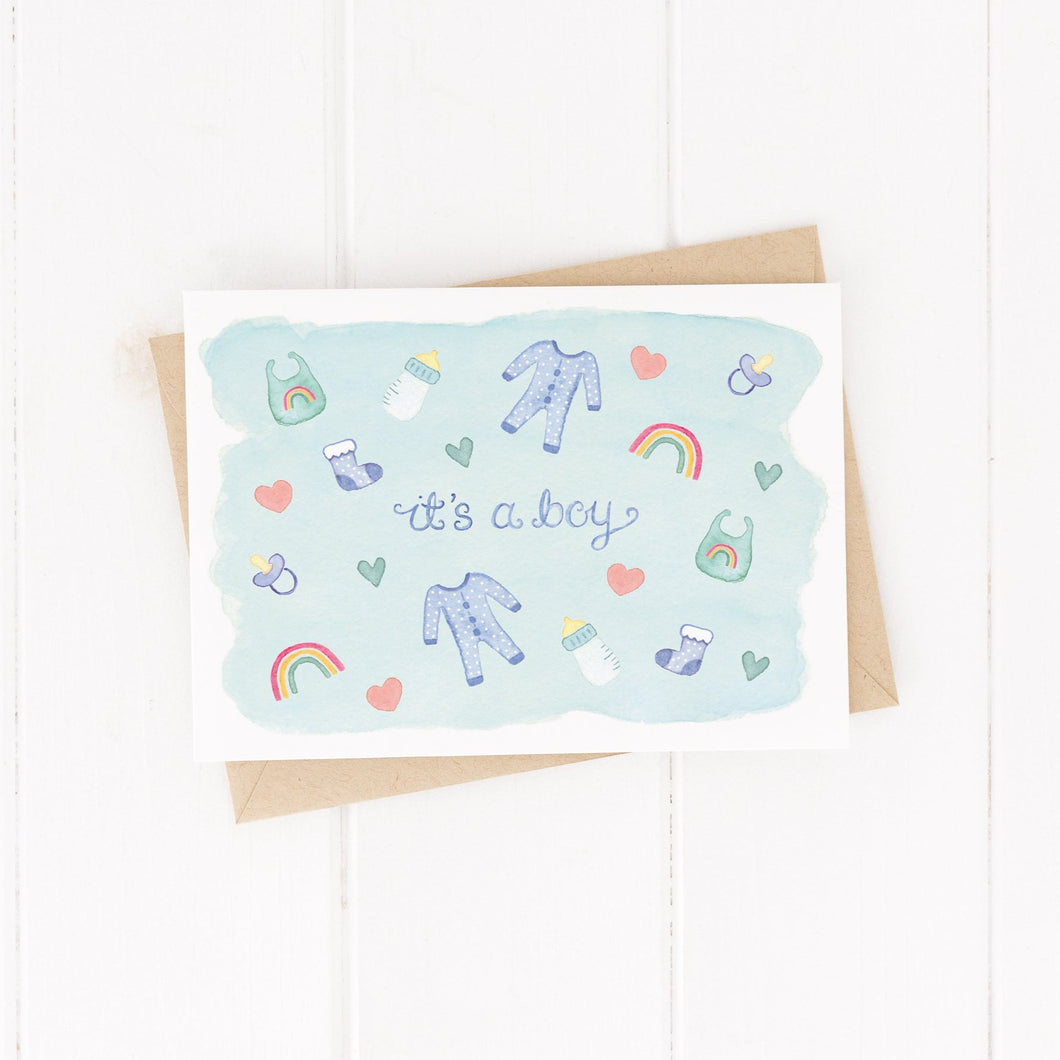 A lovely new baby card with the words, 'it's a boy' surrounded by baby item illustrations such as, baby grows, bottles, bibs, rainbows and more.