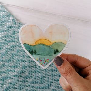 a sunset landscape watercolour illustration decal, hand painted into a heart shape to pop on your laptop, bottle or mirror