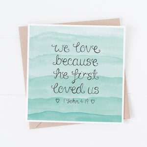 Christian love card with the words, 'we love because he first loved us' from 1 John 4 lettered onto a pretty blue watercolour background, a lovely card to celebrate a couple on their wedding day or write a love note to your spouse.