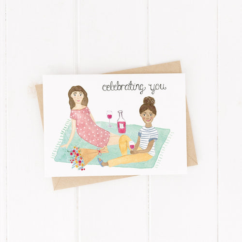 birthday card with an illustration of two girls having a picnic with a bouquet of flowers and glasses of wine with the words celebrating you hand lettered on the birthday card