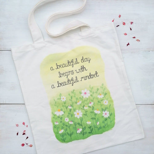 A pretty tote bag with the words, a beautiful day begins with a beautiful mindset, lettered above an illustration of daisies in a meadow.