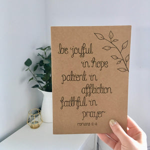 A stunning Bible verse journal with the words, 'Be joyful in hope, patient in affliction, faithful in prayer' with a dainty leaf pattern drawn above the scripture.