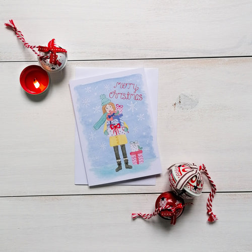 A pretty Christmas card with an illustration of a girl in the snow carrying gifts, with the words merry christmas painted in red on the design