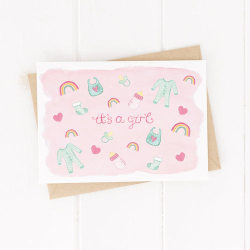 A sweet new baby girl greeting card with the words, 'It's a girl' surrounded by pink baby item illustrations such as hearts, socks, sleep suits, dummies and more.