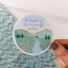 Load image into Gallery viewer, A dreamy landscape sticker with the words he leads me beside peaceful streams hand painted inside a beautiful meadow illustration, with a flowing stream and wildflowers.