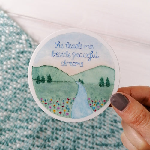 A dreamy landscape sticker with the words he leads me beside peaceful streams hand painted inside a beautiful meadow illustration, with a flowing stream and wildflowers.