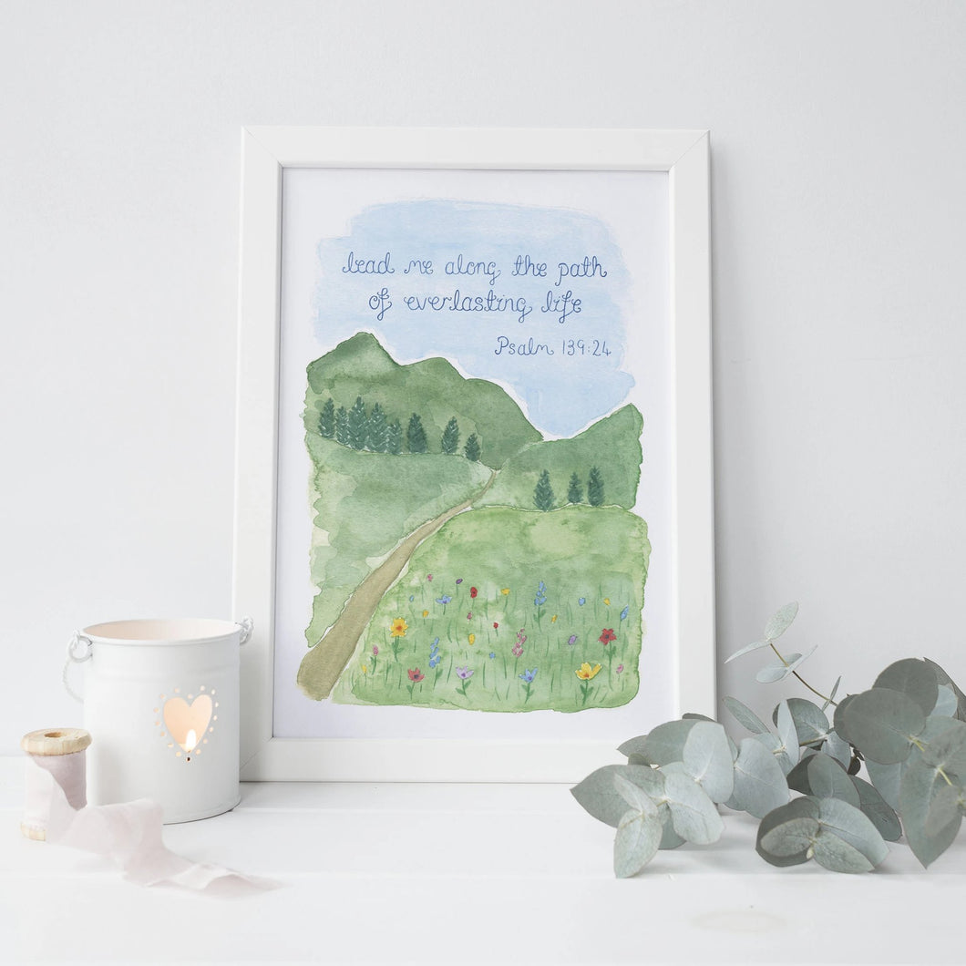 A beautiful Bible verse print with the words from Psalm 139:24, 'lead me along the path of everlasting life,' lettered onto a watercolour landscape of mountains and meadow of wildflowers with a path leading through it.