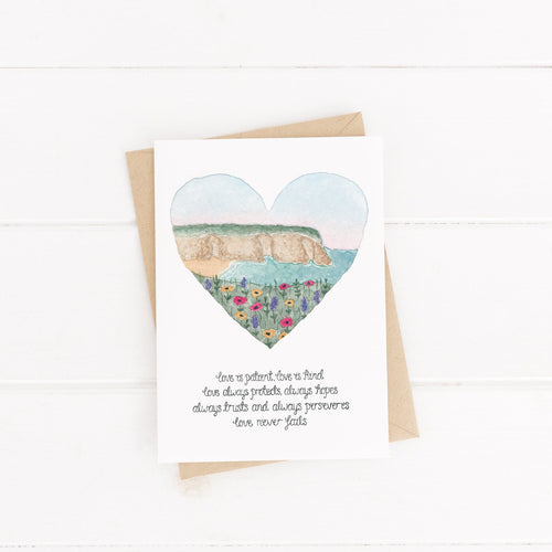 A stunning card inspired by 1 Corinthians with the words, 'love is patient, love is kind, love always protects, always hopes, always trusts and always perseveres, love never fails.' Above the verse is a landscape illustration of cliff tops and the sea painted into a heart shape.