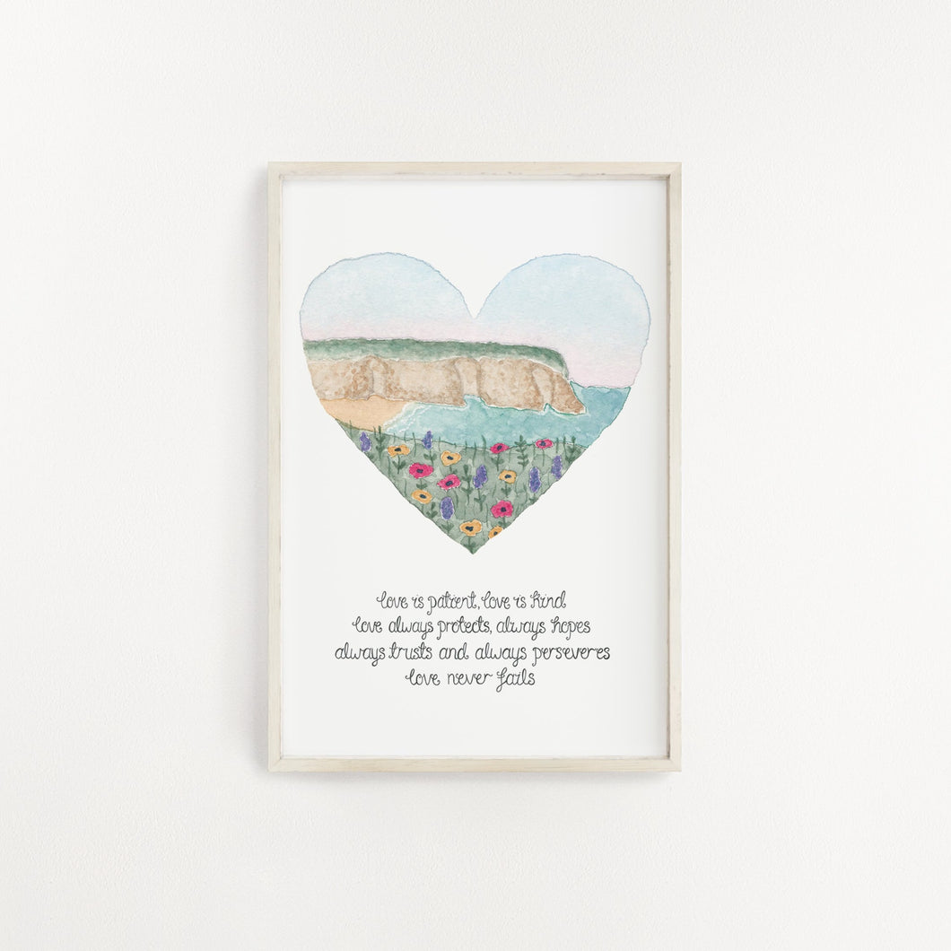 A stunning wall print inspired by 1 Corinthians with the words, 'love is patient, love is kind, love always protects, always hopes, always trusts and always perseveres, love never fails.' Above the verse is a landscape illustration of cliff tops and the sea painted into a heart shape.