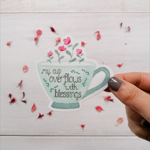 A pretty tea cup sticker with the words, my cup overflows with blessings, hand lettered inside the design with pretty pink flowers overflowing.