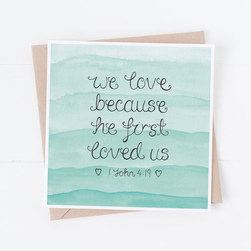 Christian love card with the words, 'we love because he first loved us' from 1 John 4 lettered onto a pretty blue watercolour background, a lovely card to celebrate a couple on their wedding day or write a love note to your spouse.