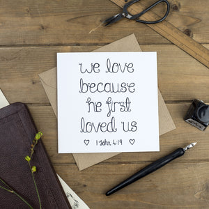 A simple yet stunning Christian wedding card with the words 'we love because He first loved us' hand lettered on the design.